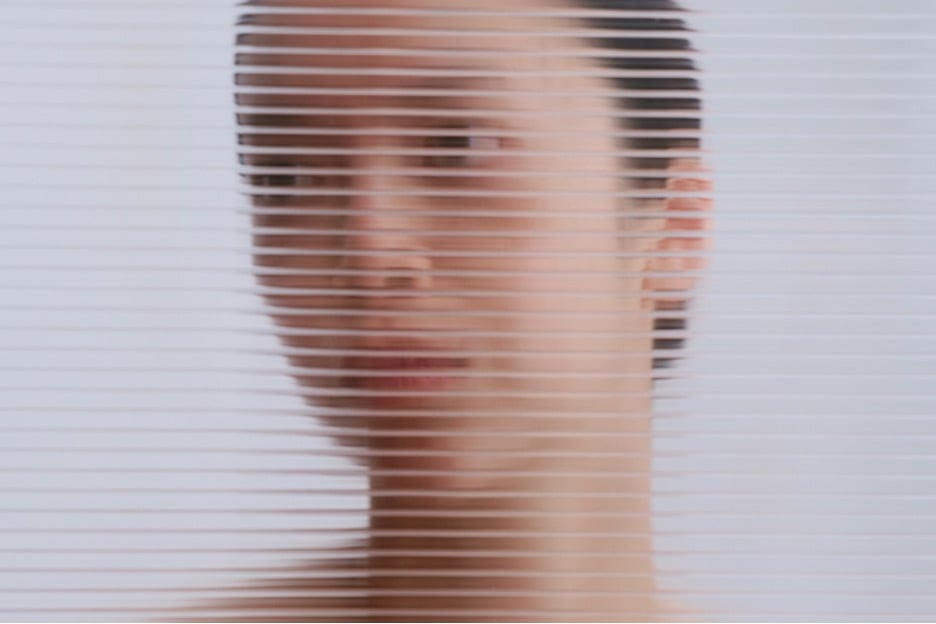 A woman gazes thoughtfully through window blinds, with a softly blurred background enhancing the sense of cognitive dissonance